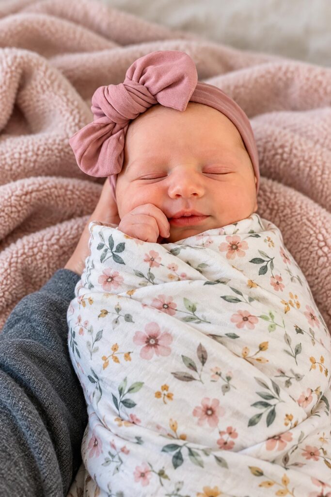 Sleeping newborn baby girl wearing a dusty pink bow headband and wrapped in a delicate floral swaddle blanket.