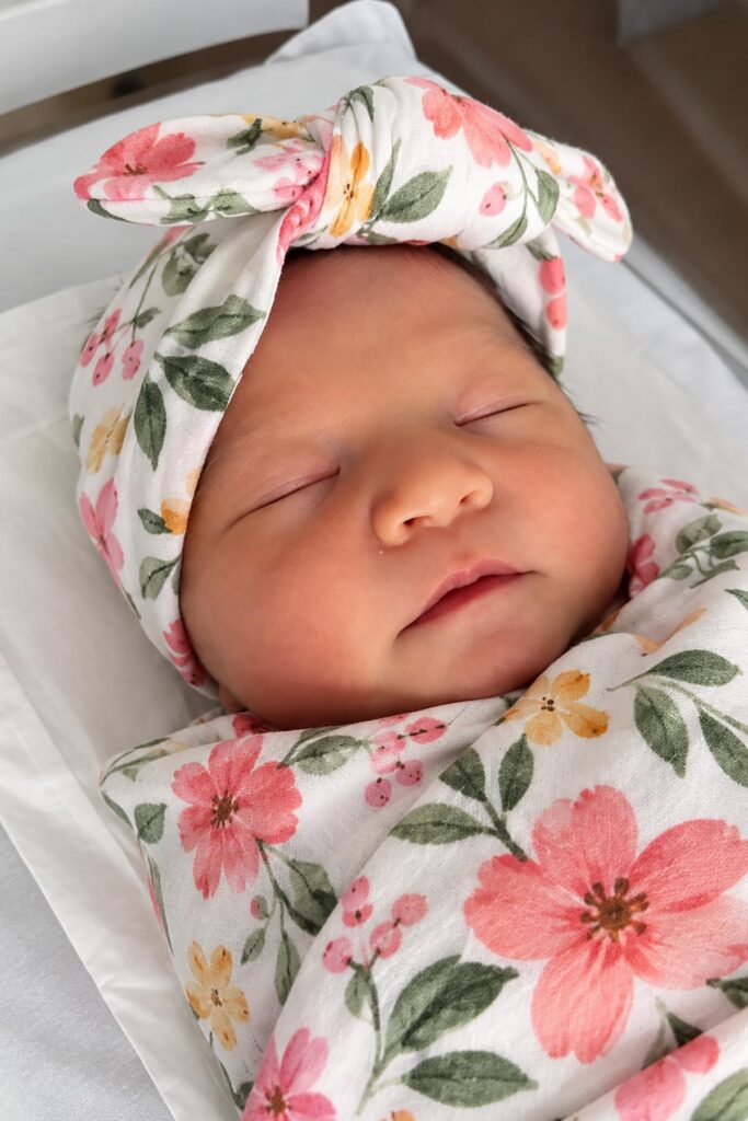 Sleeping newborn baby girl wrapped in a pink floral swaddle blanket with a matching bow headband, resting peacefully in a hospital bassinet.