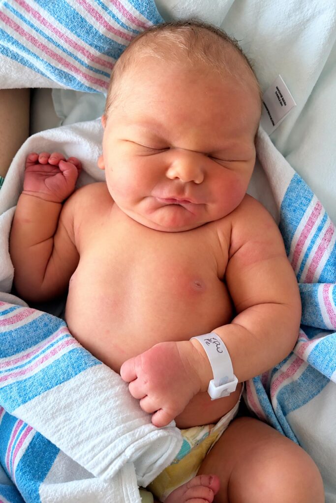 Newborn baby sleeping peacefully in a hospital blanket with tiny clenched fists, rosy cheeks, and a hospital identification bracelet.