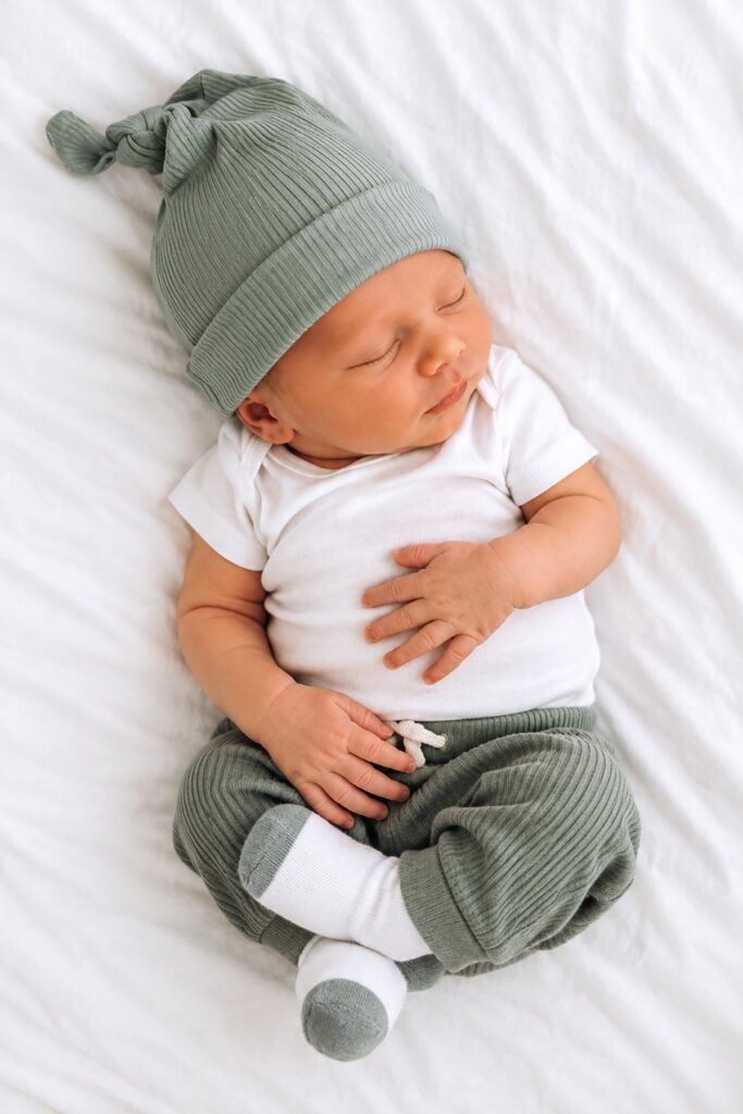 Sleeping newborn baby boy wearing a sage green knotted hat, matching pants, and white onesie while lying on a soft white blanket.