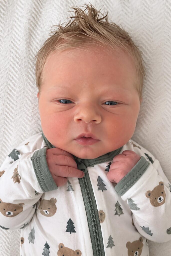 Close-up photo of a newborn baby boy with spiky blond hair wearing a white sleeper with green trim and woodland animal prints.