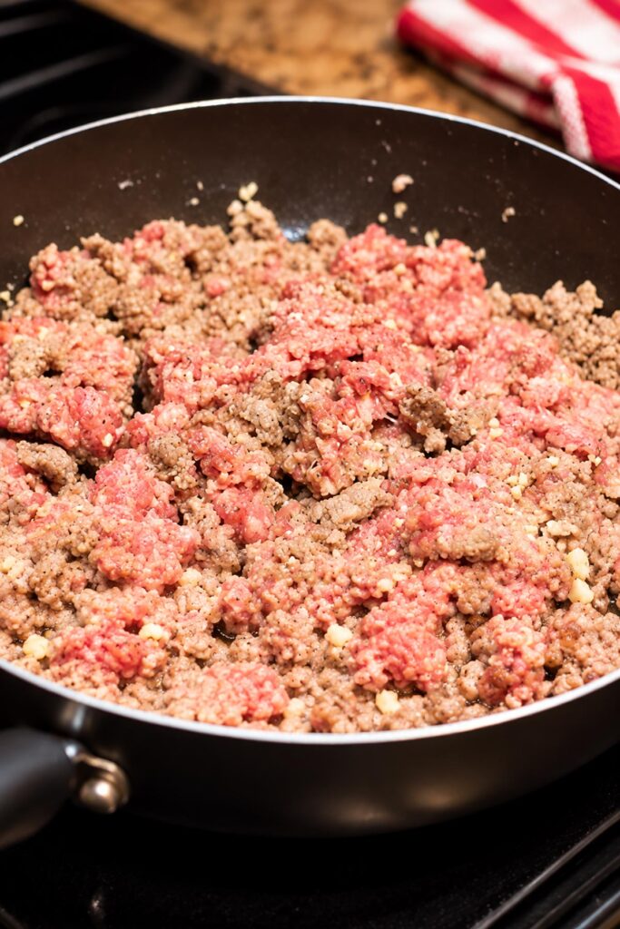Ground sausage and minced garlic cooking in a large nonstick skillet, with pink and browned meat crumbles for homemade lasagna roll-ups.