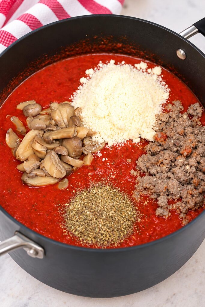 Ground sausage, sliced mushrooms, Parmesan cheese, and Italian seasoning added to tomato pasta sauce in a large pot before mixing.