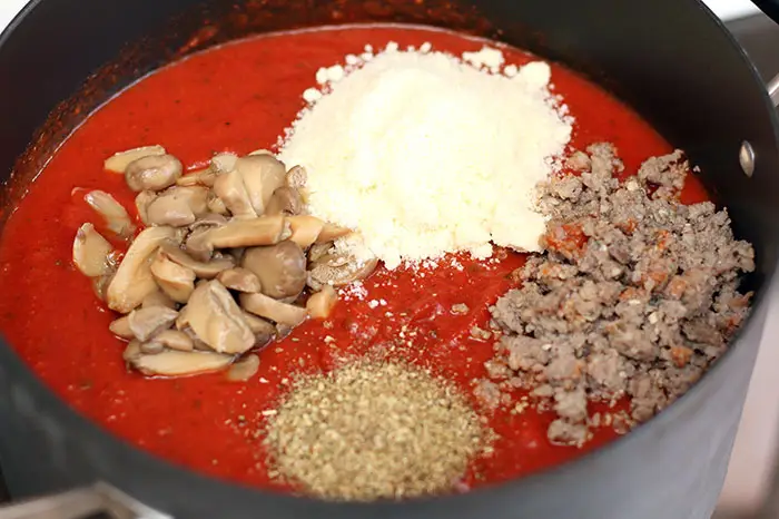 pasta sauce topped with mushrooms, parmesan, ground sausage, and oregano in a dutch oven