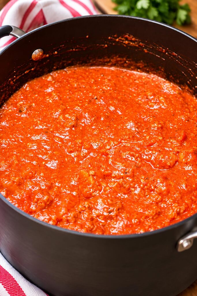 Homemade meaty pasta sauce simmering in a large black pot, featuring a rich tomato base, ground sausage, and Italian seasonings.