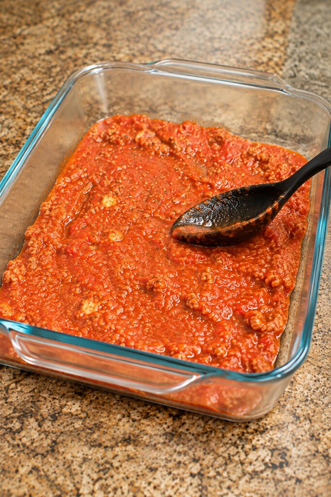 Spreading homemade meat sauce with a black spoon into a glass 9x13 baking dish to prepare cheesy lasagna roll-ups.