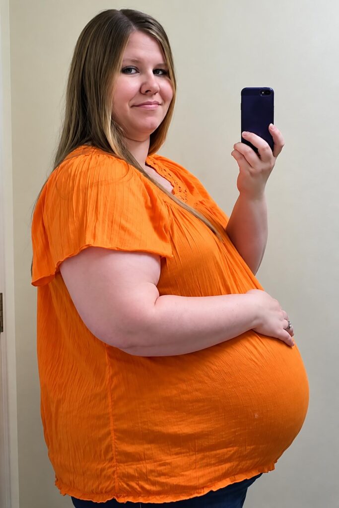 Pregnant woman in a bright orange maternity top showing her large third-trimester baby bump while taking a mirror selfie.