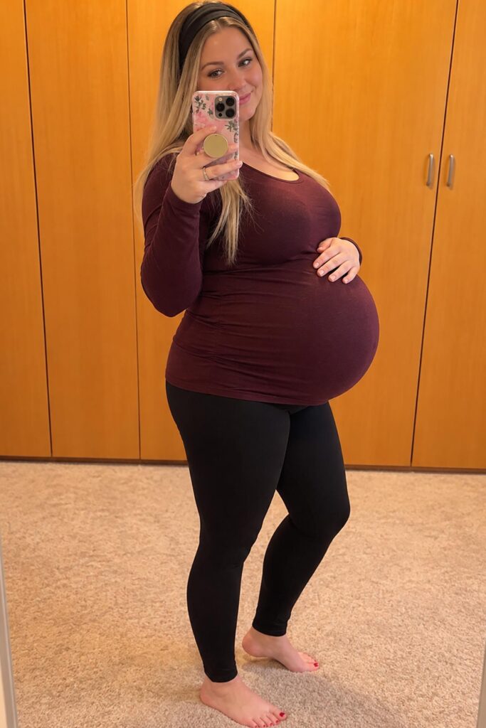 41 weeks pregnant woman showing her full-term baby bump in a burgundy maternity top and black leggings while taking a mirror selfie.