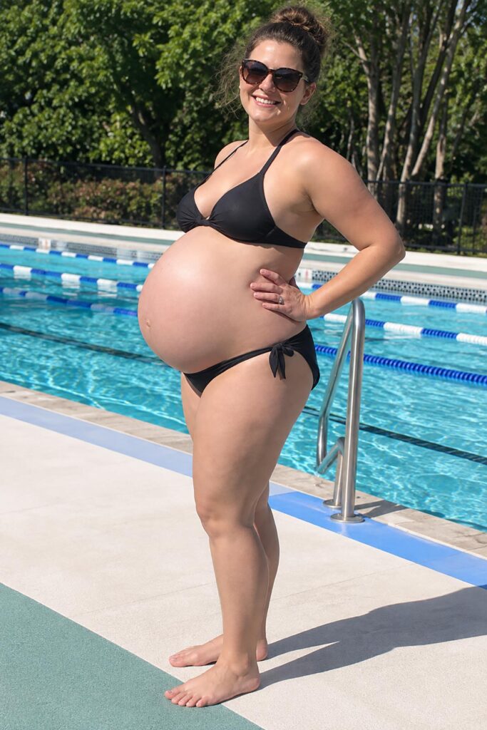 Woman at 42 weeks pregnant showing her large baby bump in a black bikini while standing beside a swimming pool on a sunny day.