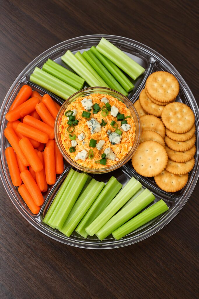 Homemade buffalo chicken dip served on a party platter with baby carrots, celery sticks, crackers, and blue cheese crumbles.