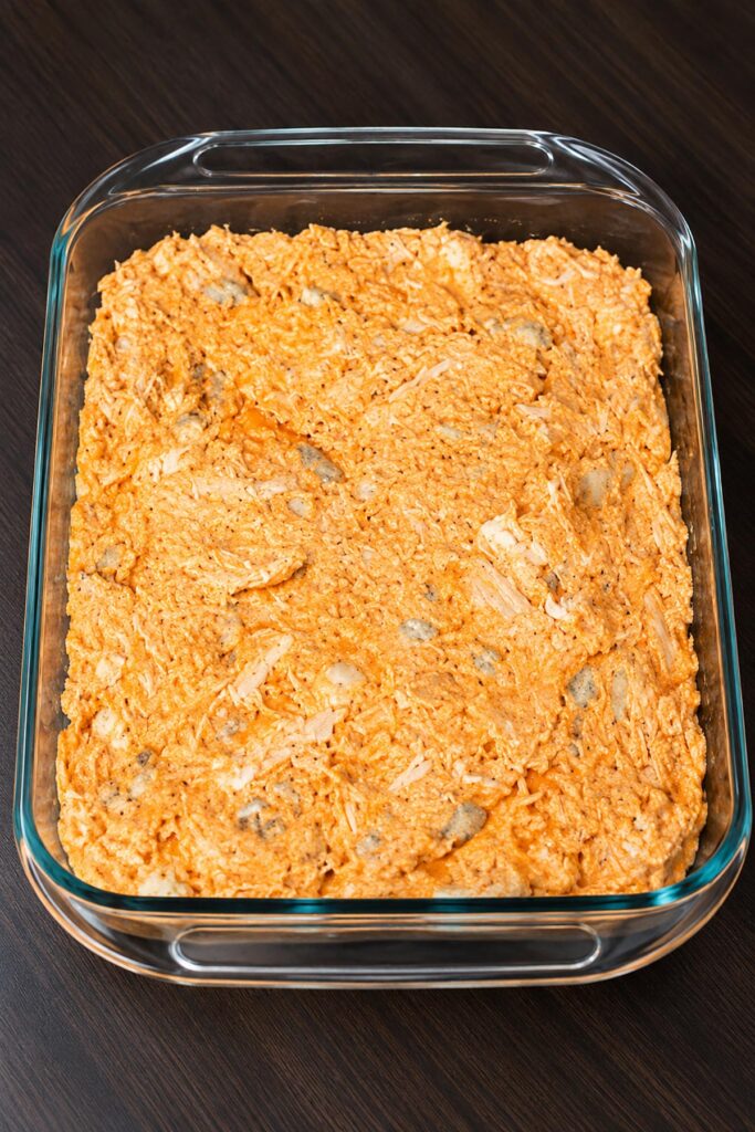 Unbaked buffalo chicken dip mixture with shredded chicken, cheese, and buffalo sauce spread in a glass casserole dish.
