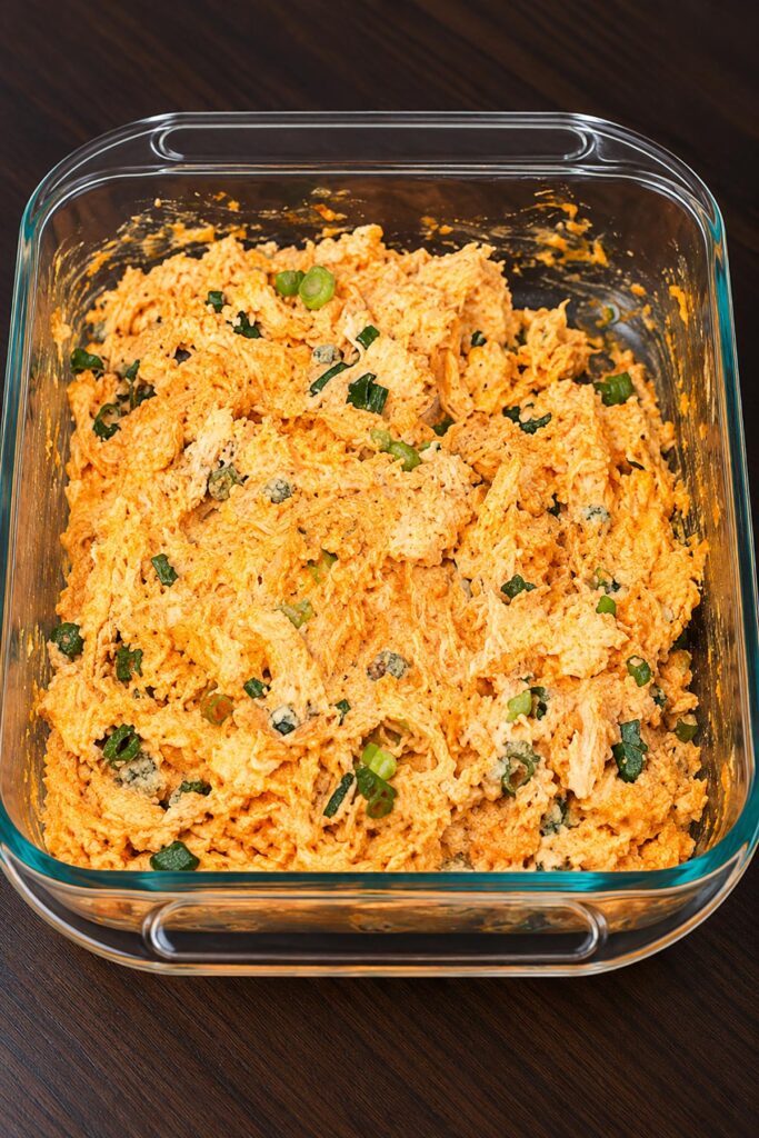 Creamy buffalo chicken dip mixture with shredded chicken, melted cheese, and chopped green onions in a glass baking dish.