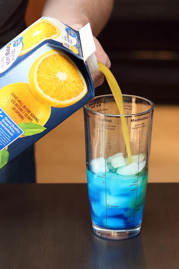 pouring orange juice into cocktail shaker with blue liquid