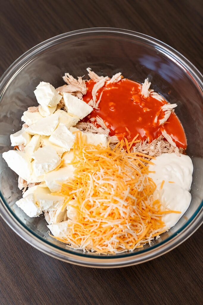 Buffalo chicken dip ingredients in a glass bowl, including shredded chicken, cream cheese, buffalo sauce, dressing, and shredded cheese.