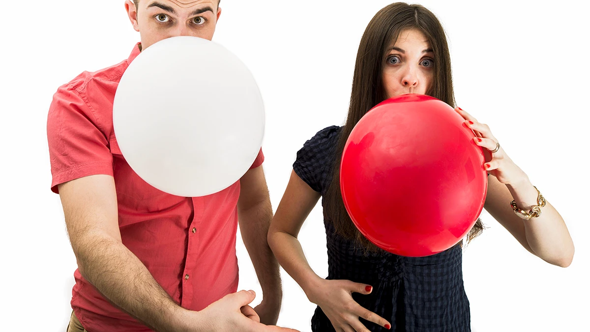 Couple blowing up white and red balloons for a funny pregnancy announcement photo against a white background.