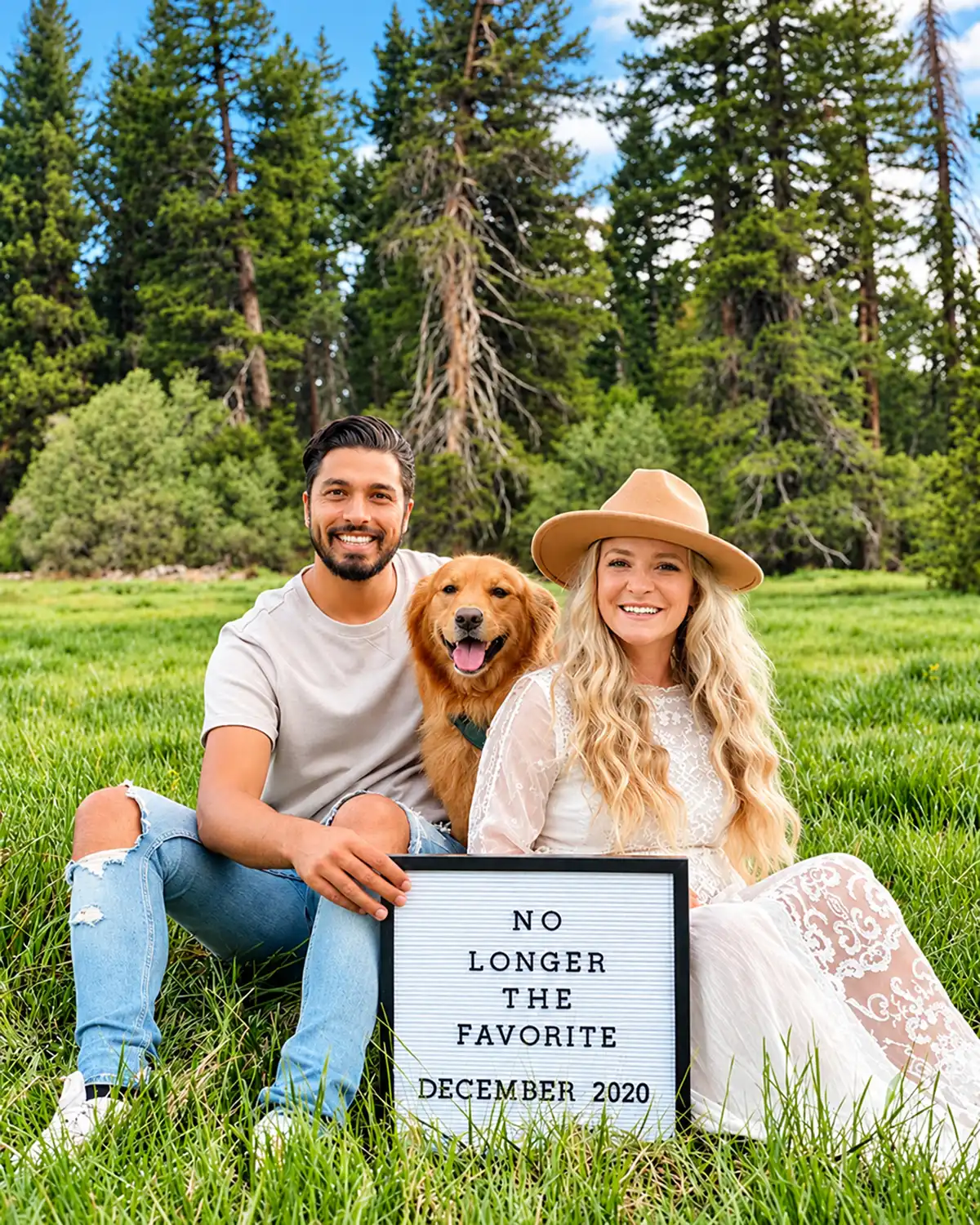 Expectant couple with golden retriever holding a No Longer the Favorite pregnancy announcement sign in a grassy meadow.