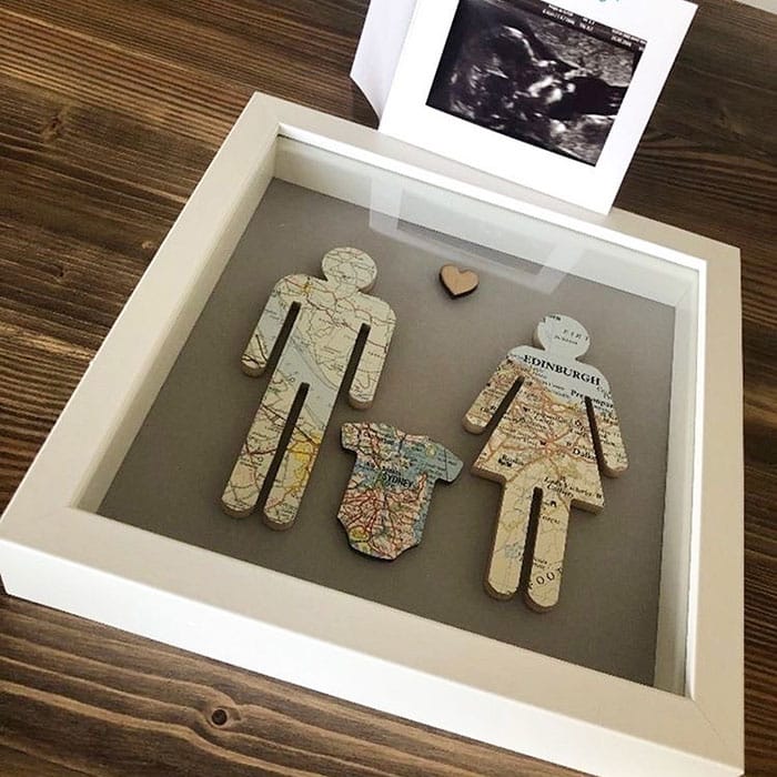 a shadowbox frame of silhouettes for a man, woman, and baby bodysuit, each created out a paper map