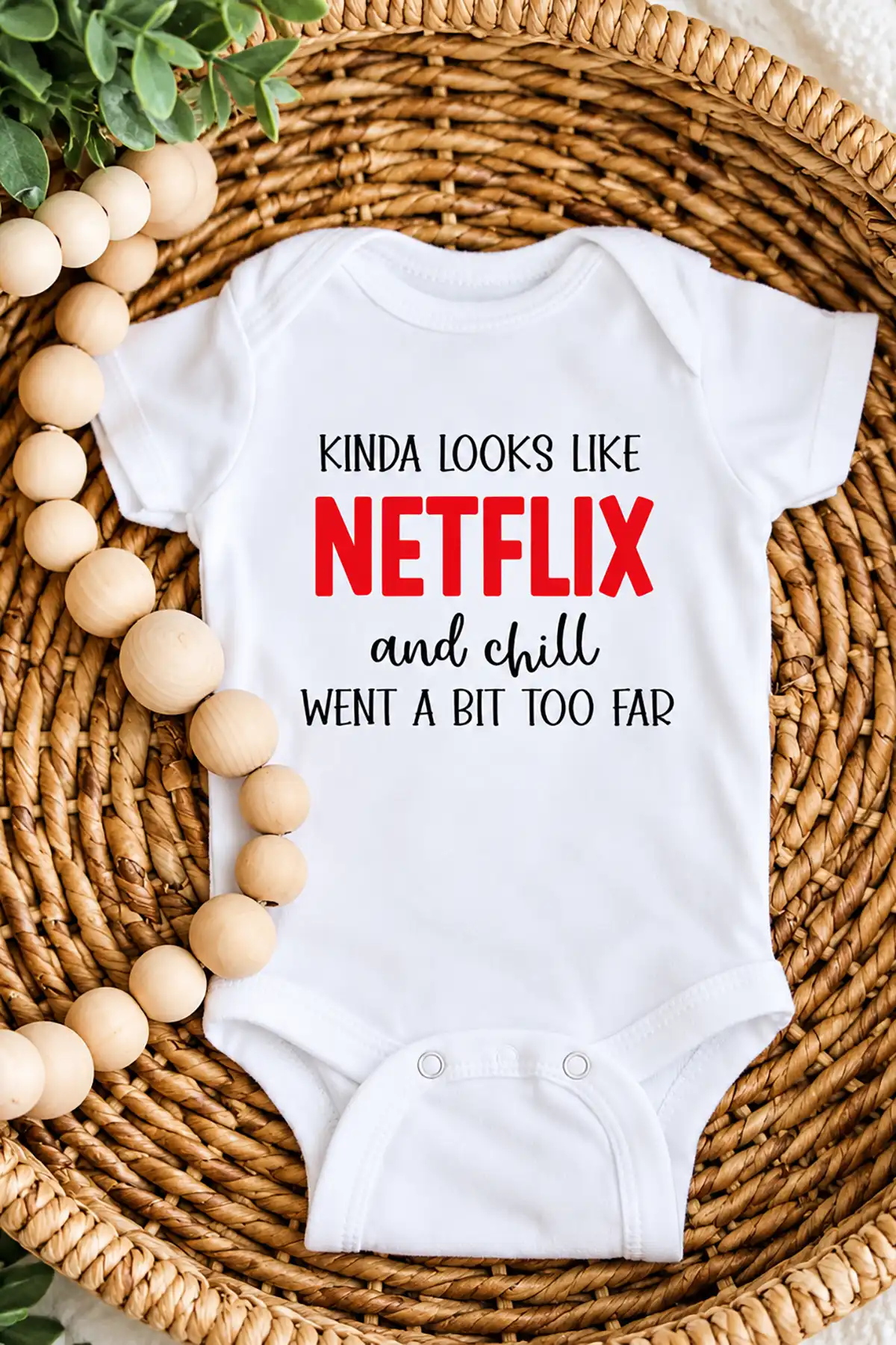 White baby onesie reading Kinda Looks Like Netflix and Chill Went a Bit Too Far on a woven basket with wooden beads.