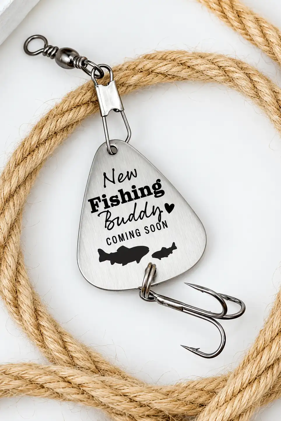 Silver fishing lure engraved New Fishing Buddy Coming Soon with fish silhouettes, surrounded by rustic rope.
