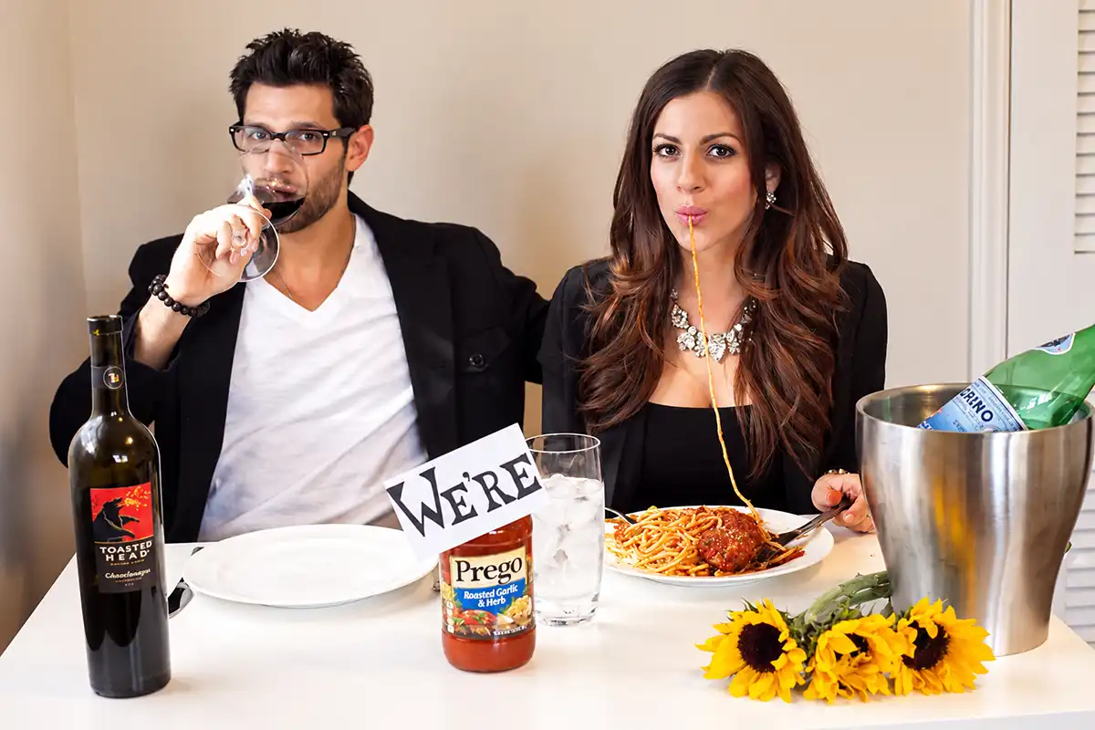 Couple announcing pregnancy with a Prego pasta sauce jar, spaghetti dinner, and wine for a funny baby reveal photo.