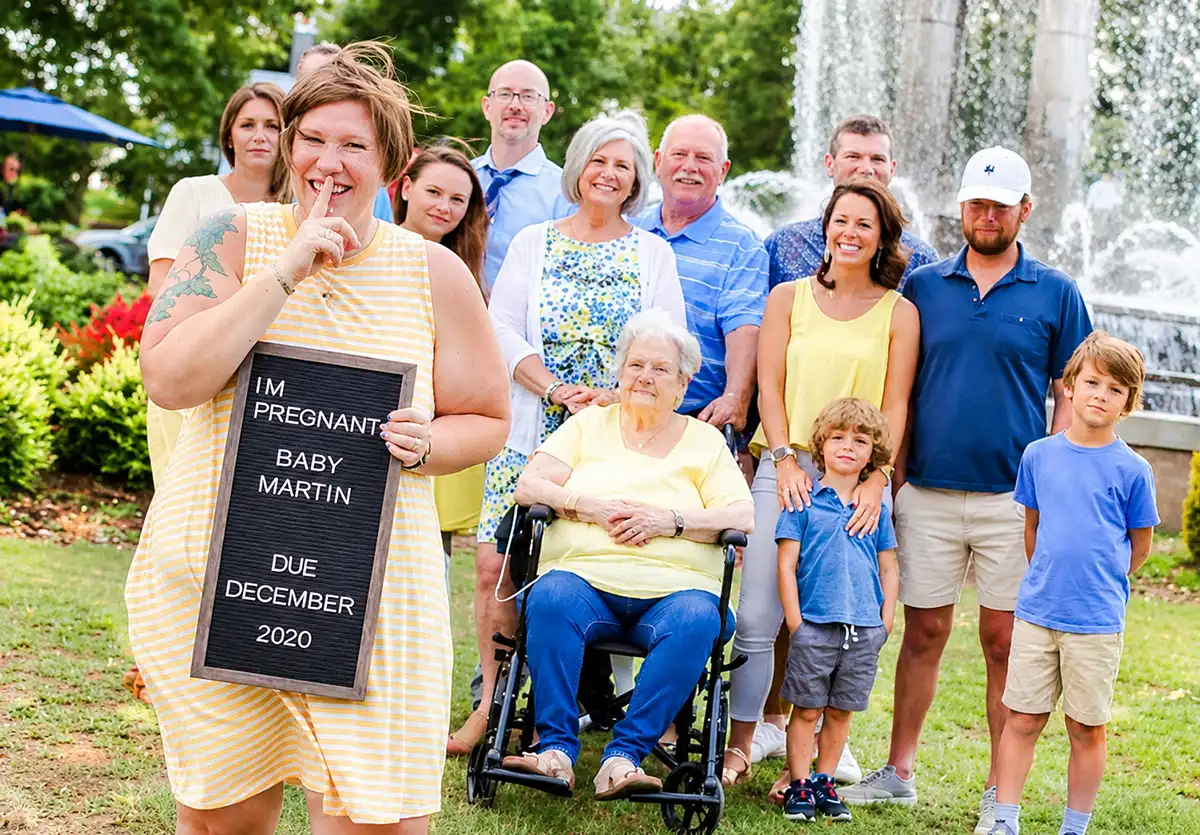 Expectant mother holding an I'm Pregnant letterboard in front of extended family for a surprise pregnancy announcement photo.