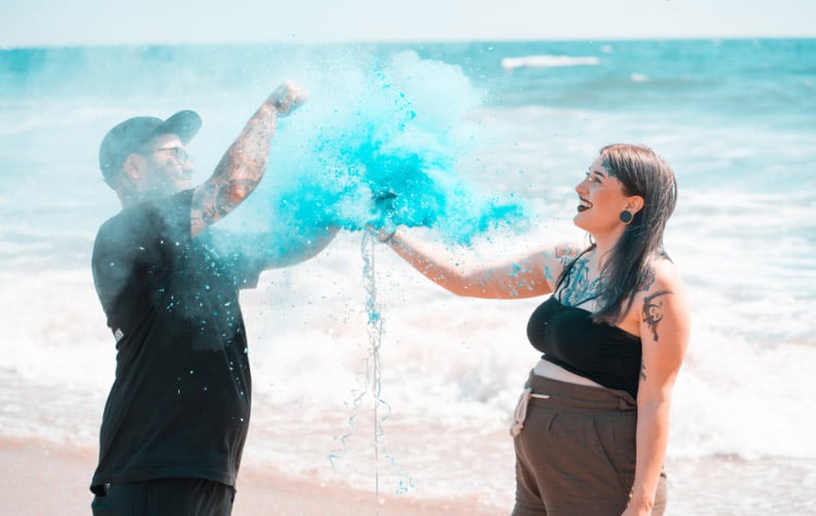 pregnancy woman and partner standing on the beach at the moment a black gender reveal balloon is popped to reveal blue powder smoke for a boy