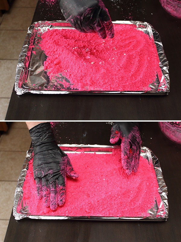 pink cornstarch being spread on a baking sheet with large chunks broken up by gloved hands