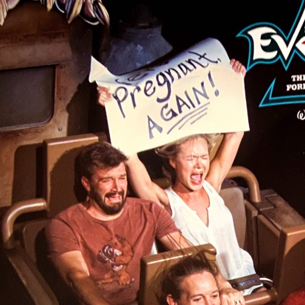woman holding up a sign reading "we're pregnant again" while riding a rollercoaster