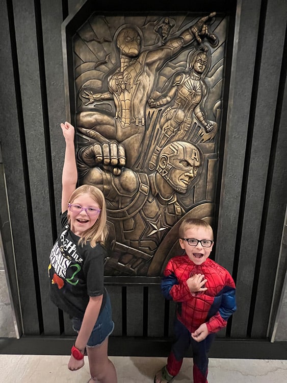 two children, one dressed as Spider-Man, standing in front of a bronze Marvel art piece