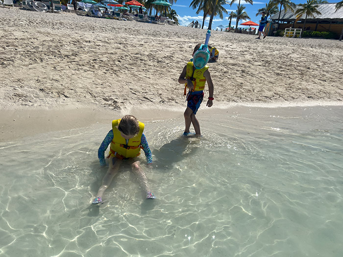 two children in shallow water at the beach, one sitting and the other standing with a full-face snorkel mask on