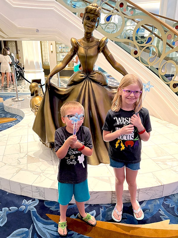 two children holding star wands in front of the bronze Cinderella statue on the Disney Wish