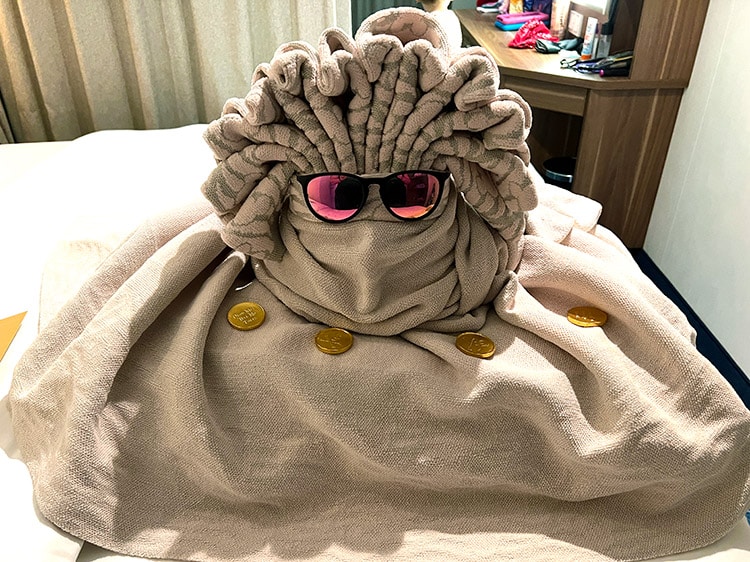 an unidentifiable towel animal wearing sunglasses with chocolate pirate coins around it on a bed inside a Disney Wish stateroom