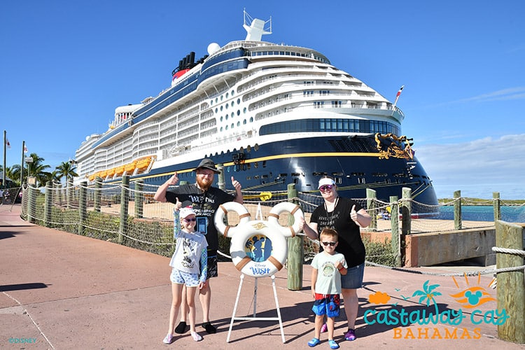 a family of four posing with Mickey-shaped lifesaver rings in front of the Disney Wish cruise ship