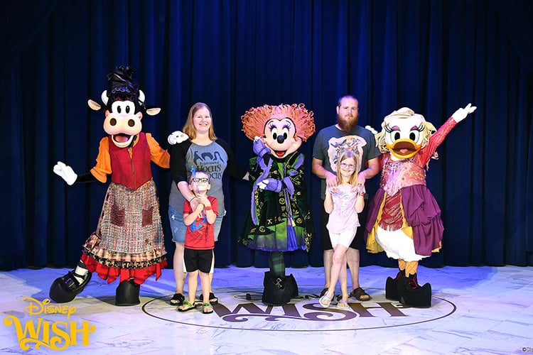 a family posing with Minnie, Daisy, and Clarabelle dressed as the Sanderson sisters on the Disney Wish Grand Hall stage