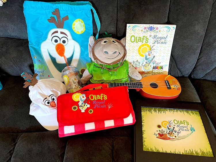 all of the gifts children receive at Olaf's Royal Picnic, including a bag, troll plush, hat, picnic blanket, activity book, mandolin, necklace, drinking cup, and photo