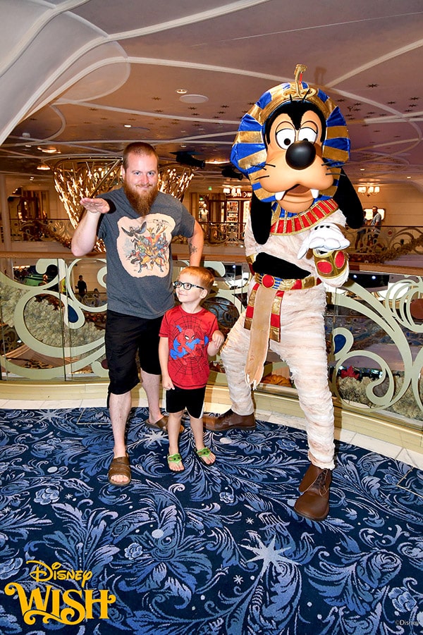 a father and son posing with Goofy dressed as a mummy on the Disney Wish