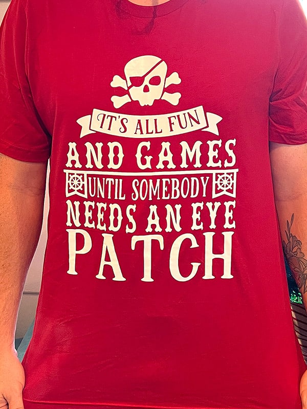 red shirt with a skull and crossbones wearing an eye patch and the words "It's all fun and games until somebody needs an eye patch"