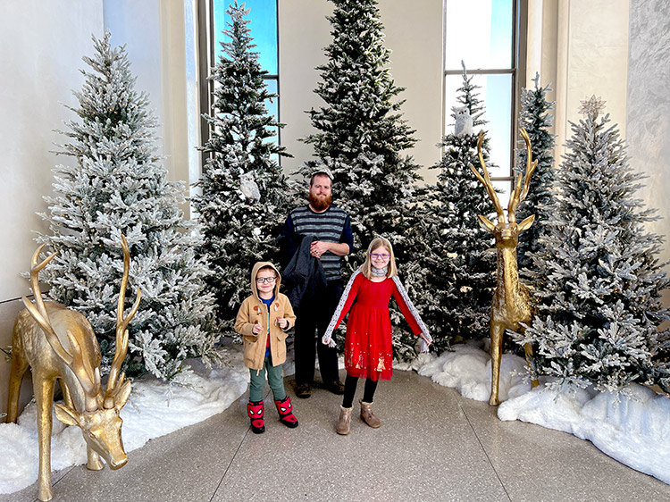 a father, son, and daughter posing with gold reindeer and Christmas trees in a winter display at Top of the Rock in NYC