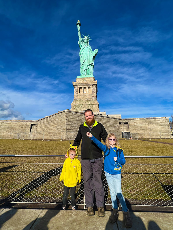 a father, son, and daughter posing in front of the Statue of Liberty