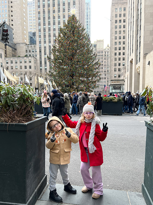 a boy and girl posing in front of the Rockefeller Christmas tree during the day