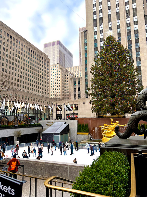 a wide view of the Rockefeller center at Christmas time featuring the giant Christmas tree, the Prometheus statue, and The Rink with people ice skating