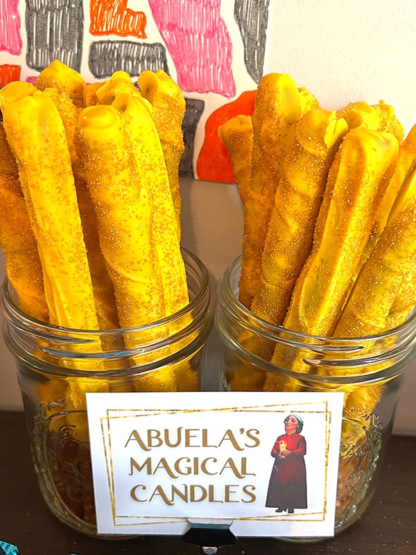 two Ball canning jars holding giant pretzel sticks covered in yellow chocolate and gold sugar glitter with a card featuring Abuela Alma from Encanto and the text "Abuela's magical candles"