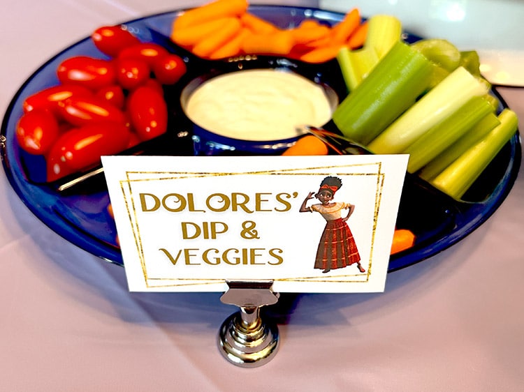 a veggie tray with tomatoes, baby carrots, celery sticks, and ranch with a card featuring Dolores from Encanto and the text "Dolores' dip & veggies"