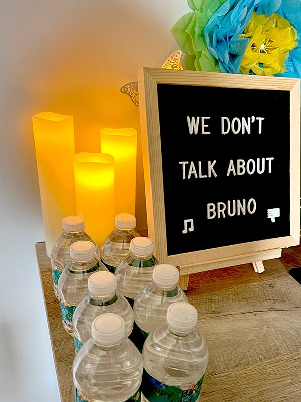 closeup of part of a table featuring three glowing LED candles of various lengths, small water bottles, and a letter board that says "We don't talk about Bruno"