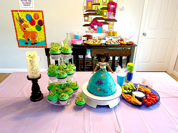a wide shot of an Encanto birthday party arrangement with cake, cupcakes, a candle, and veggie tray on a large table with more food on a server behind it and a Casita house made from cardstock on the wall
