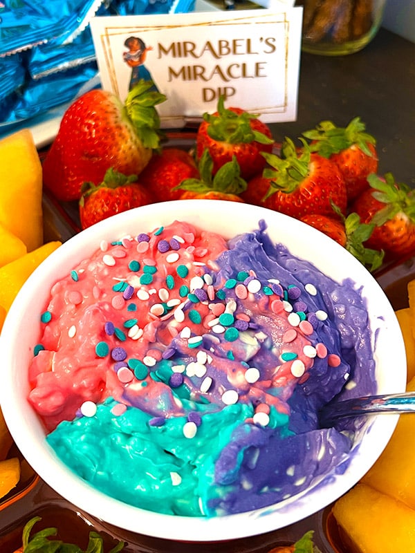 bowl of creamy fruit dip in pink, teal, and purple colors and sprinkles on top on a tray of cantaloupe and strawberries; in the background is a card featuring Mirabel from Encanto with text reading "Mirabel's miracle dip"