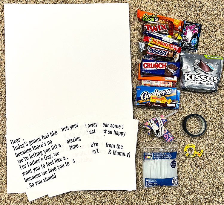 flat lay of supplies needed to make a Father's Day candy bar poster, including foam board, a printed message, candy bars, hot glue gun and glue sticks, masking tape, and double-sided tape