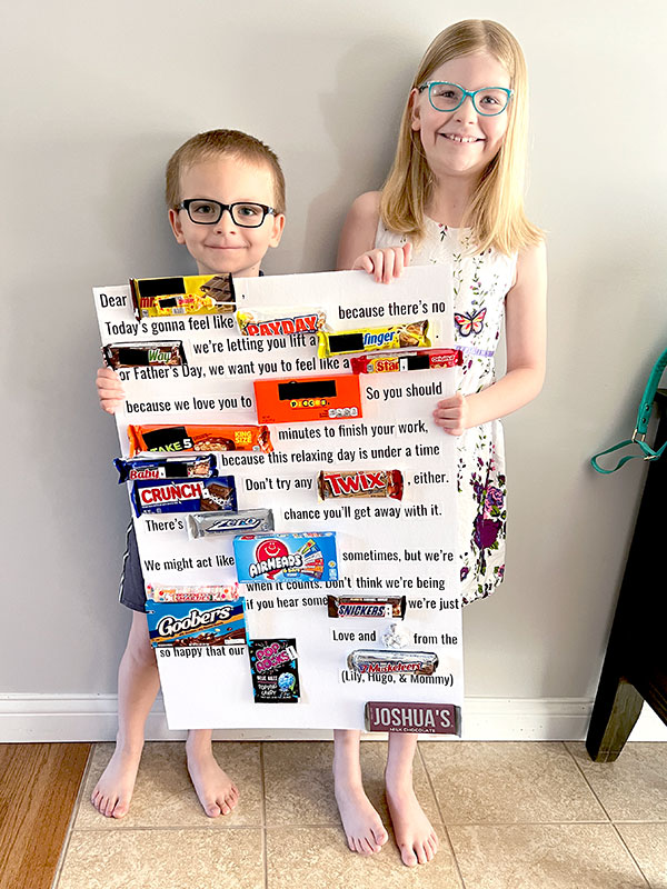 two young children holding up a large white foam board that has a Father's Day message with various types of candy filling in for the words their names contain
