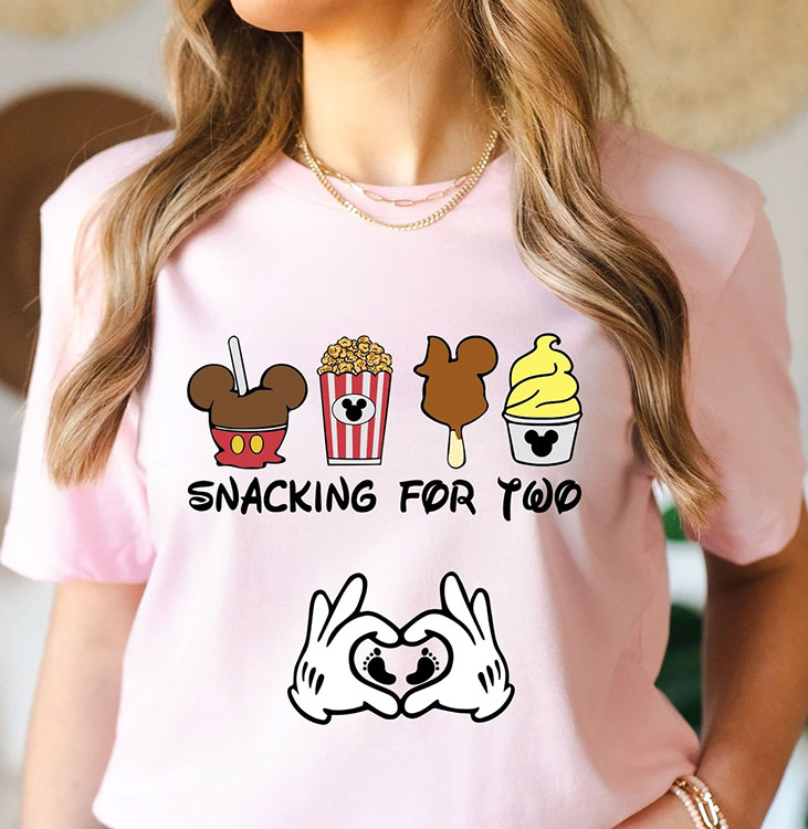 woman wearing pink t-shirt featuring several Disney park snacks, gloved Mickey Mouse in a heart shape around baby footprints, and the text "Snacking for two"