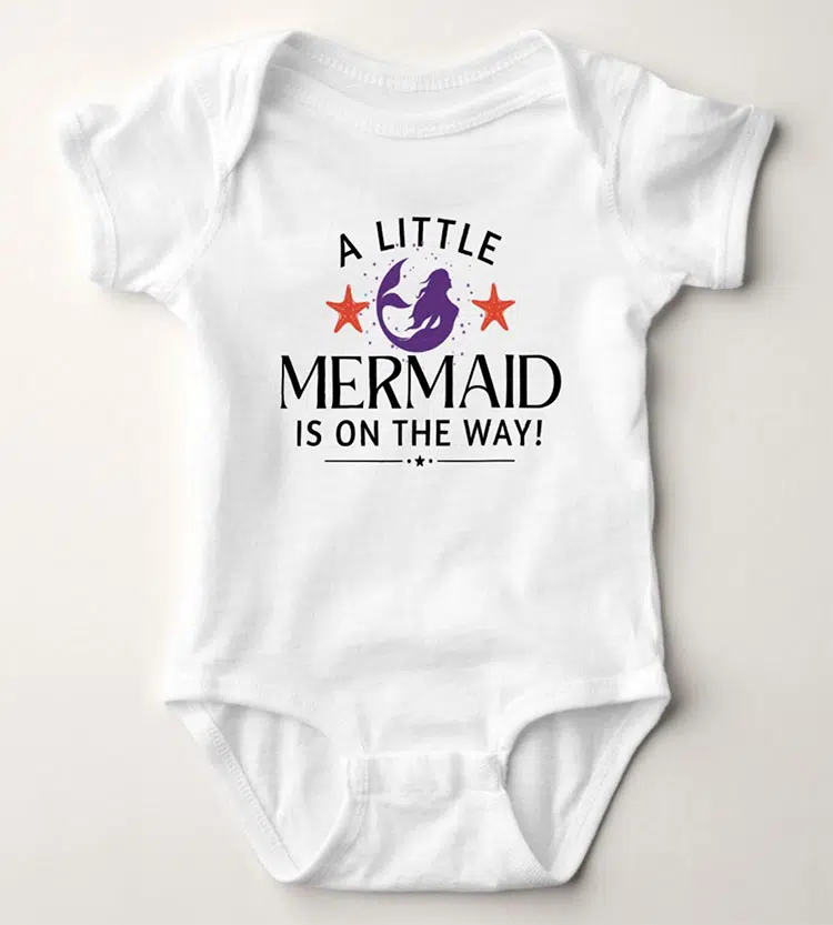 a white baby bodysuit featuring a purple mermaid and pink stars and black text reading "A little mermaid is on the way!"